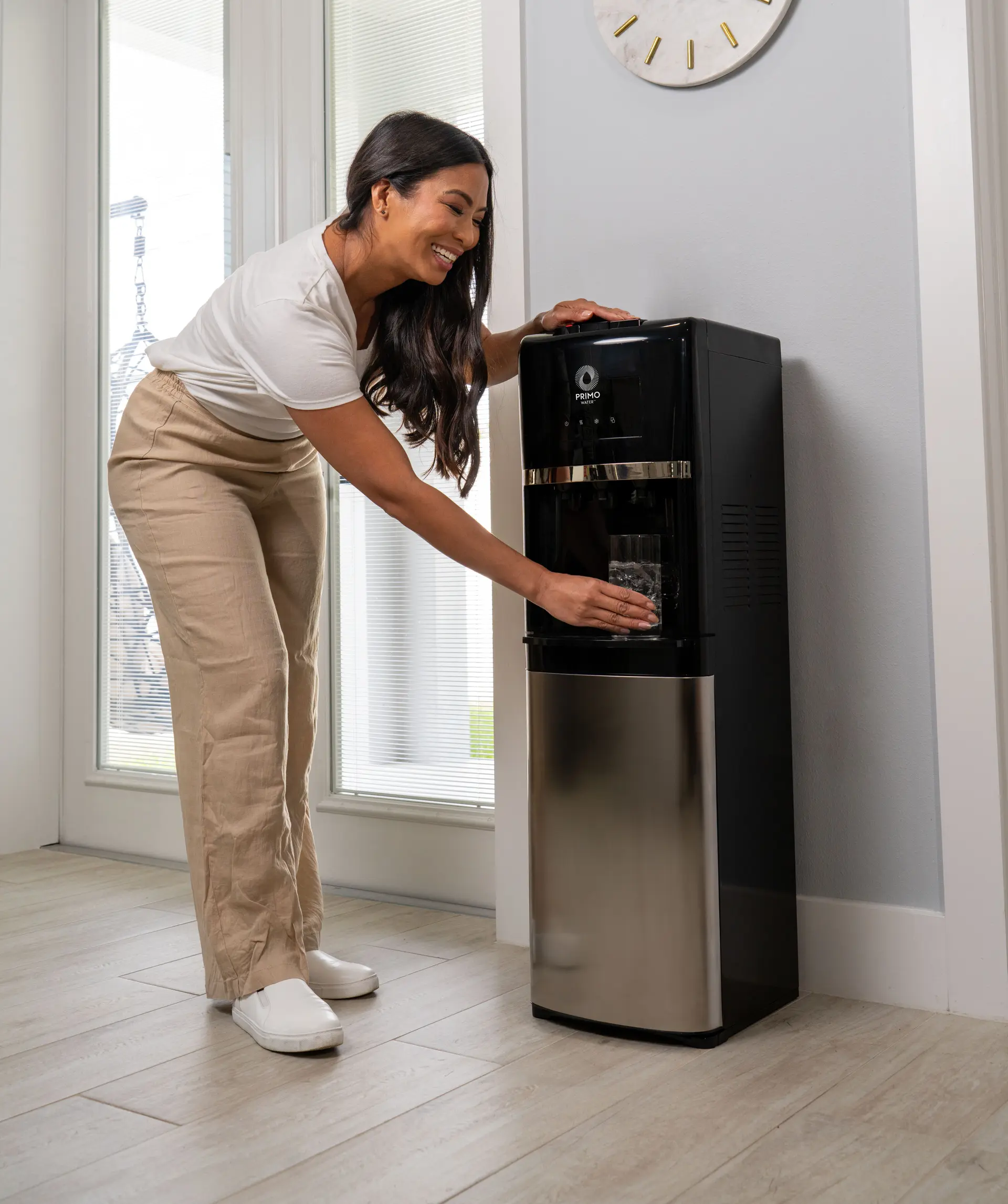 Woman in beige pants and white shirt smiling while filling a glass with water from a Primo water dispenser.