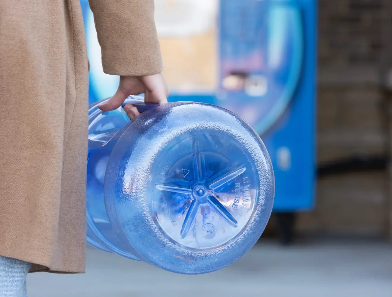 Person holding a Primo Water® water jug by the handle, wearing a brown coat walking up to an exchange station.