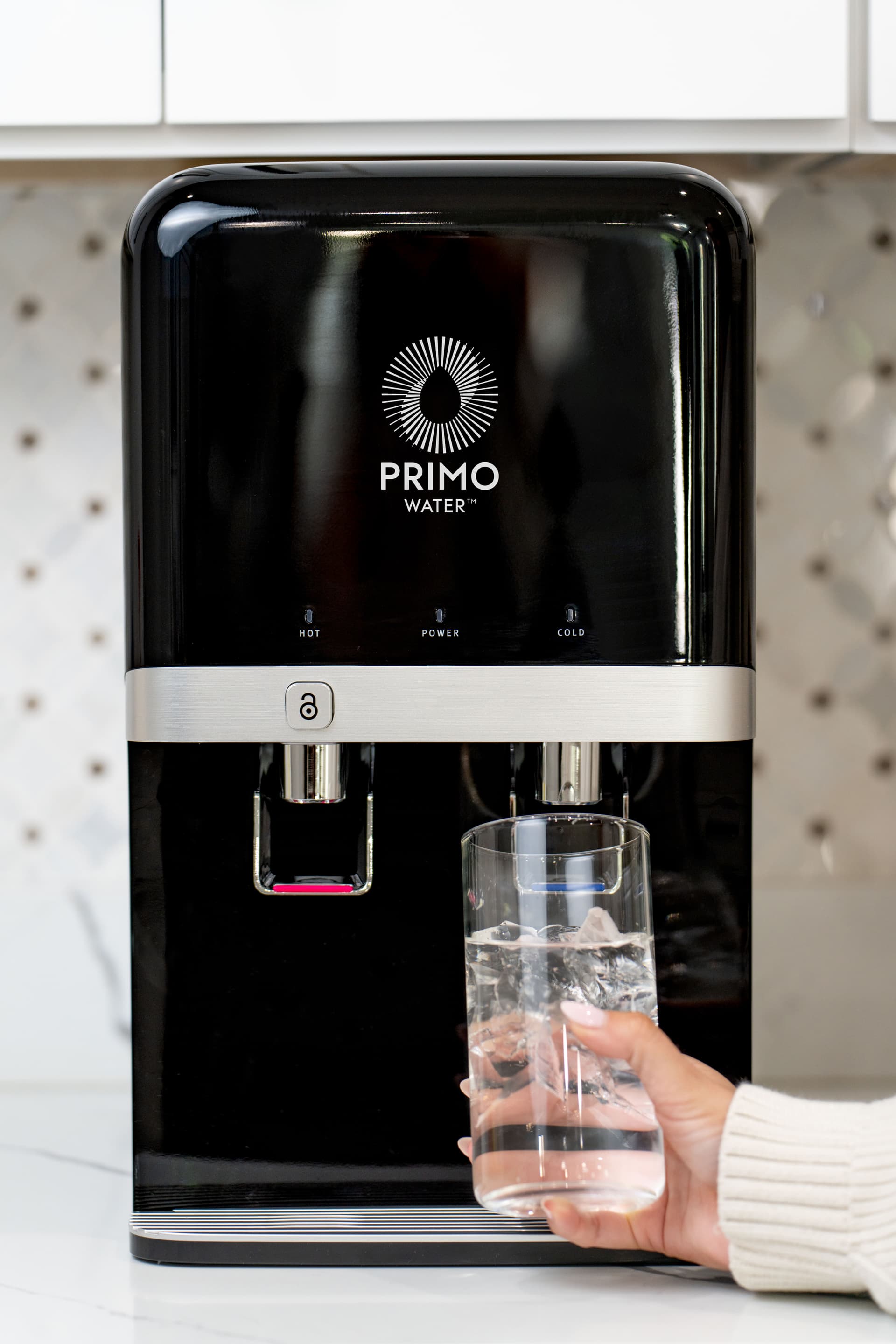 A person fills a glass with water from a black countertop Primo water dispenser featuring hot and cold options.