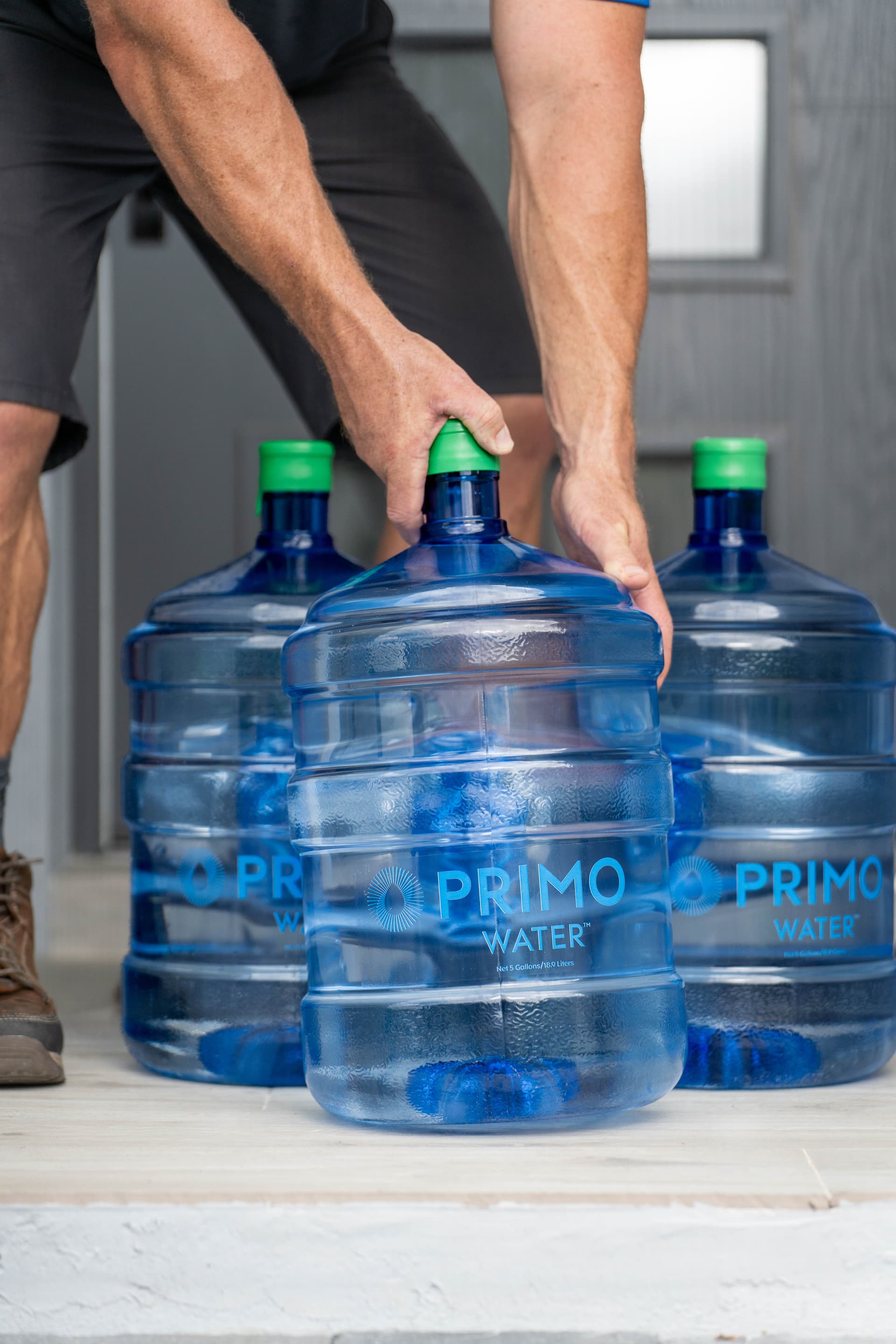 Person lifting a large blue Primo Water® water jug after delivery, with two more jugs on the floor.